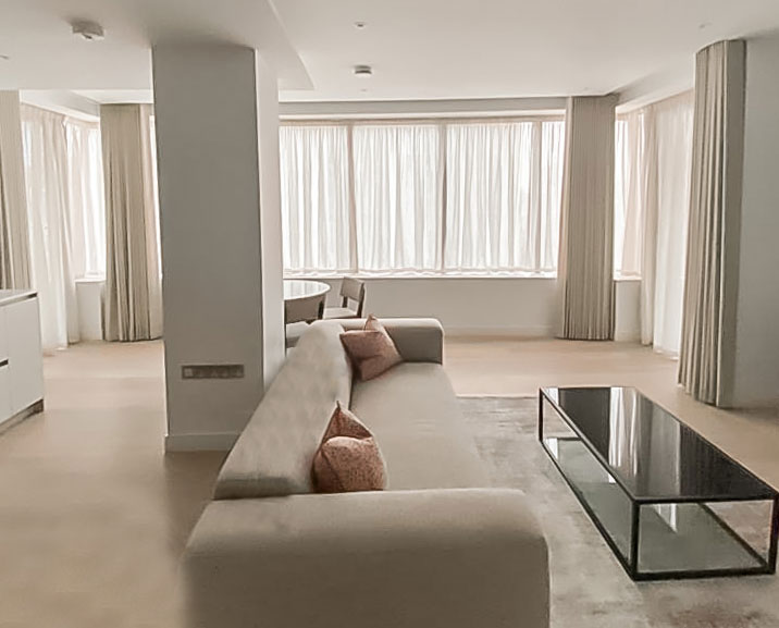 In a large living room the windows have net curtains and neatly pleated floor length curtains